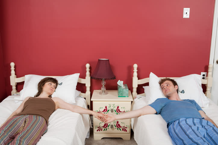 Una pareja acostada en dos camas individuales separadas extendiendo sus manos para agarrárselas