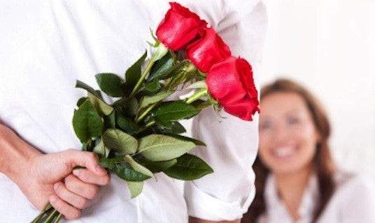 Un hombre sorprende a su pareja con un ramo de rosas rojas, símbolo clásico de amor y romance, capturando un momento de felicidad y anticipación.